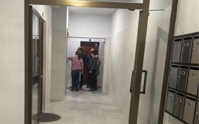 Entrada reformada de un portal comunitario, con suelos y paredes en tonos claros, una puerta corredera de vidrio y varios buzones a la derecha. Al fondo, tres personas conversan cerca del ascensor.