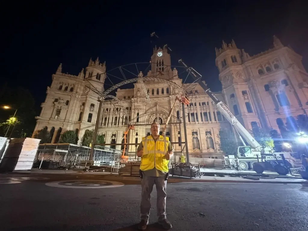 Project Manager con chaleco reflectante amarillo posa en primer plano durante la noche frente al Palacio de Cibeles en Madrid, donde se realiza un montaje con grúas y estructuras metálicas.