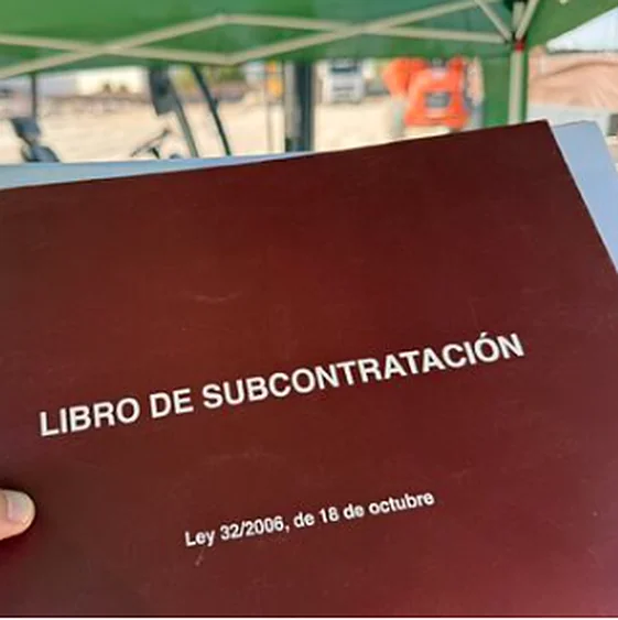 Libro de Subcontratación de la Ley 32/2006 sostenido por un trabajador en una obra, con una carpa y maquinaria al fondo.